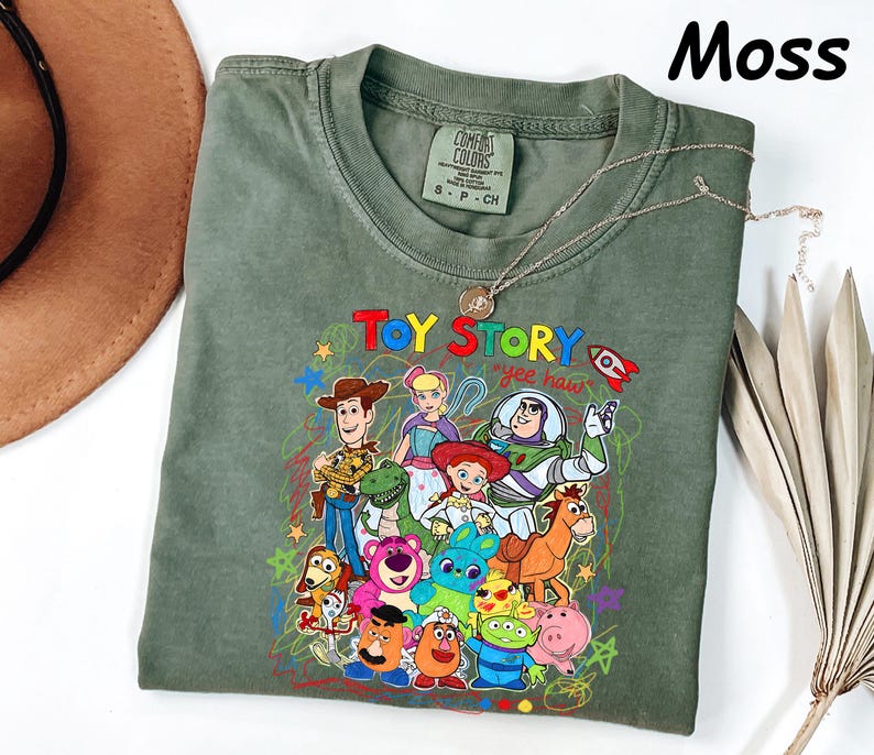 May include: Moss green t-shirt featuring a colorful graphic of Toy Story characters. The design includes Woody, Buzz Lightyear, and other beloved characters from the animated film. The words "Toy Story" and "yee haw" are also visible.