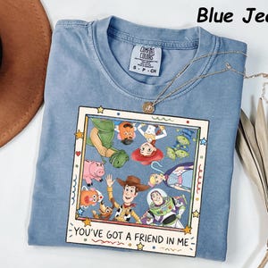 May include: A blue jean colored t-shirt featuring a graphic print of Toy Story characters inside a square frame. The text "YOU'VE GOT A FRIEND IN ME" is printed at the bottom of the frame. The shirt has a crew neckline and a Comfort Colors brand label.