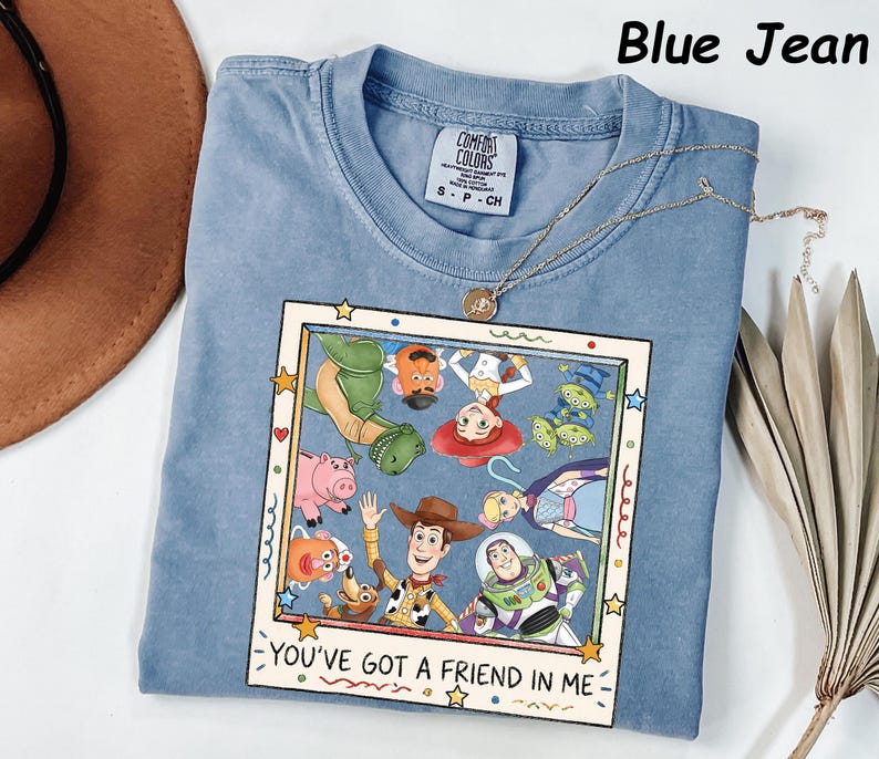 May include: A blue jean colored t-shirt featuring a graphic print of Toy Story characters inside a square frame. The text "YOU'VE GOT A FRIEND IN ME" is printed at the bottom of the frame. The shirt has a crew neckline and a Comfort Colors brand label.