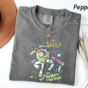 May include: A gray Comfort Colors t-shirt featuring a graphic of Buzz Lightyear with the text "Buzz Lightyear" and "To Infinity and Beyond." The shirt has a crew neck and short sleeves. A silver necklace with a pendant is on the shirt.