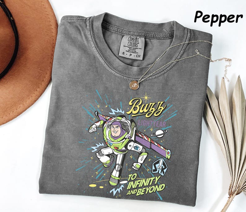 May include: A gray Comfort Colors t-shirt featuring a graphic of Buzz Lightyear with the text "Buzz Lightyear" and "To Infinity and Beyond." The shirt has a crew neck and short sleeves. A silver necklace with a pendant is on the shirt.