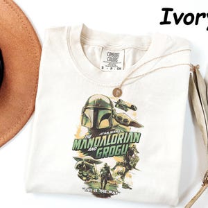 Peut inclure: T-shirt ivoire avec un graphisme de The Mandalorian et Grogu de Star Wars. Le design comprend le casque du Mandalorien, Grogu, et le texte "THE MANDALORIAN AND GROGU" et "THIS IS THE WAY". Un collier en or est drap&eacute; sur le t-shirt.