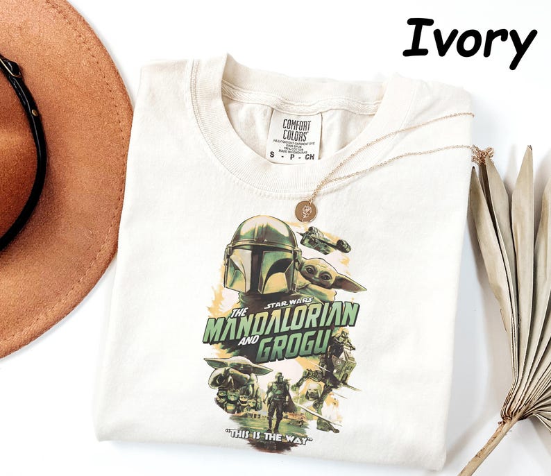 Peut inclure: T-shirt ivoire avec un graphisme de The Mandalorian et Grogu de Star Wars. Le design comprend le casque du Mandalorien, Grogu, et le texte "THE MANDALORIAN AND GROGU" et "THIS IS THE WAY". Un collier en or est drap&eacute; sur le t-shirt.