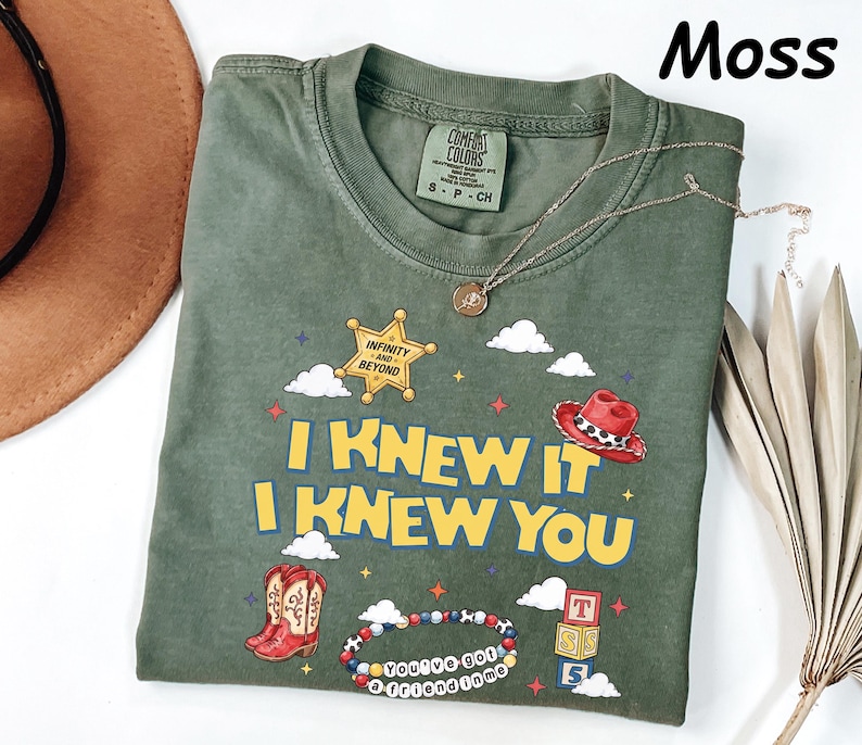 May include: A moss green short-sleeve t-shirt featuring a graphic print with the text I KNEW IT I KNEW YOU, a yellow sheriff star badge with the text INFINITY AND BEYOND, a red cowboy hat, red cowboy boots, alphabet blocks, and a beaded bracelet.