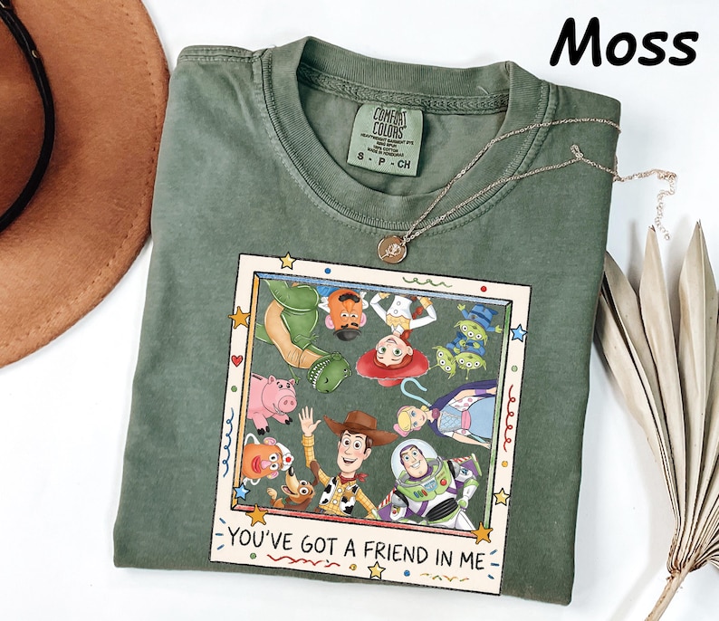 May include: A moss green crew neck t-shirt featuring a graphic print of various animated characters inside a square frame. The text "YOU'VE GOT A FRIEND IN ME" is printed at the bottom of the frame. The shirt has a Comfort Colors brand label.
