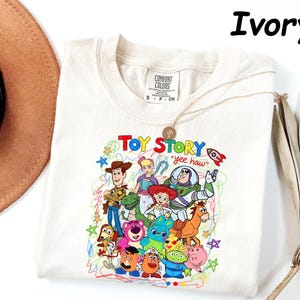 May include: Ivory-colored t-shirt featuring a colorful graphic of Toy Story characters. The design includes Woody, Buzz Lightyear, and other characters from the animated film. The text "Toy Story" and "yee haw" are also visible. The shirt is a Comfort Colors brand.