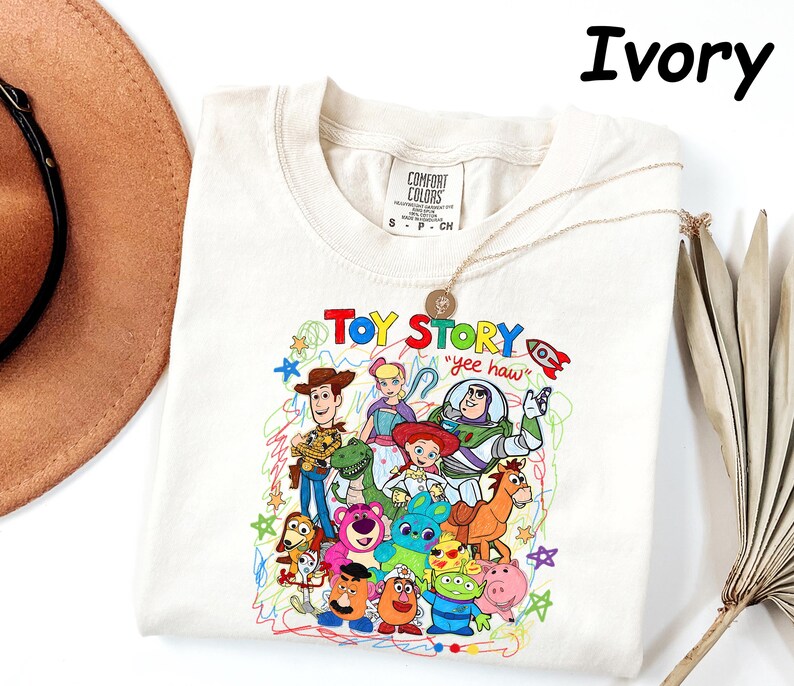 May include: Ivory-colored t-shirt featuring a colorful graphic of Toy Story characters. The design includes Woody, Buzz Lightyear, and other characters from the animated film. The text "Toy Story" and "yee haw" are also visible. The shirt is a Comfort Colors brand.