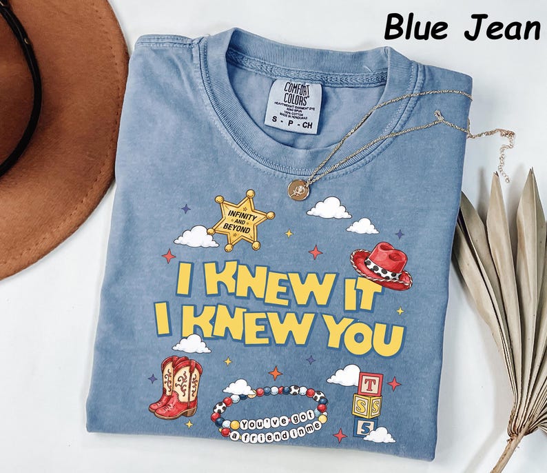 May include: A blue jean colored t-shirt featuring a graphic print with a yellow sheriff star labeled Infinity and Beyond, a red cowboy hat, red cowboy boots, a beaded bracelet with the text You've Got a Friend in Me, and blocks with letters.
