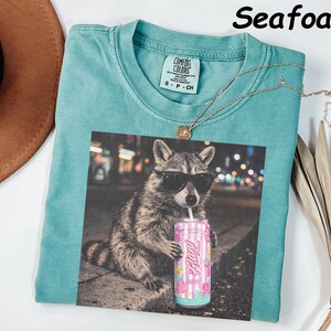 May include: Seafoam green t-shirt with a graphic of a raccoon wearing sunglasses and holding a pink can. The shirt has a Comfort Colors tag and a gold necklace. The word "Seafoam" is printed at the top.