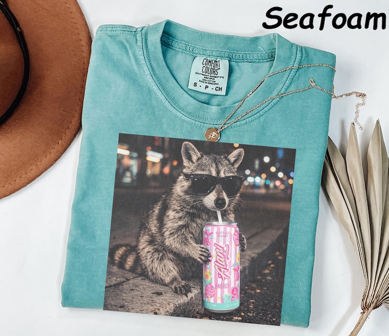 May include: Seafoam green t-shirt with a graphic of a raccoon wearing sunglasses and holding a pink can. The shirt has a Comfort Colors tag and a gold necklace. The word "Seafoam" is printed at the top.
