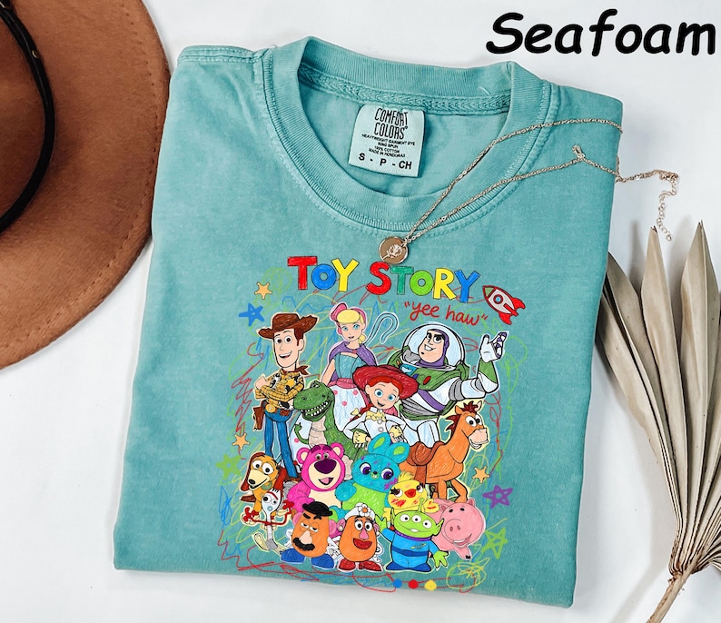 May include: Seafoam green t-shirt featuring a colorful graphic of Toy Story characters. The shirt has the words "Toy Story" and "yee haw" printed on it. A gold necklace with a pendant is draped over the shirt.