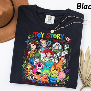 May include: Black crew neck sweatshirt featuring a colorful graphic of Toy Story characters. The design includes Woody, Buzz Lightyear, and other characters from the animated film. A gold necklace is draped across the sweatshirt.