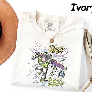 May include: Ivory-colored t-shirt featuring a graphic of Buzz Lightyear from Toy Story. The design includes the text "Buzz Lightyear" and "To Infinity and Beyond" in a comic book style. A gold necklace is draped across the shirt.