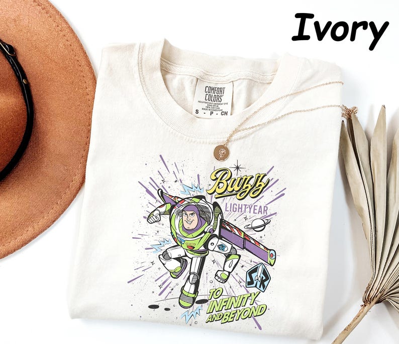 May include: Ivory-colored t-shirt featuring a graphic of Buzz Lightyear from Toy Story. The design includes the text "Buzz Lightyear" and "To Infinity and Beyond" in a comic book style. A gold necklace is draped across the shirt.