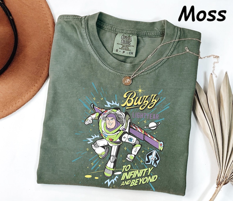 May include: Moss green t-shirt featuring a graphic of Buzz Lightyear with the text "Buzz Lightyear" and "To Infinity and Beyond." The shirt has a crew neck and short sleeves. A gold necklace is draped across the shirt.