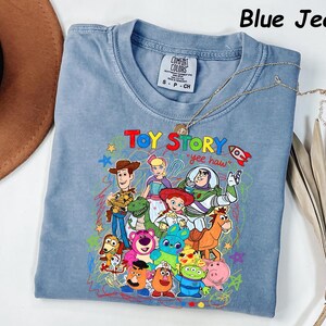 May include: Blue jean colored t-shirt featuring a colorful graphic of Toy Story characters. The text "Toy Story" and "yee haw" are displayed on the front. The shirt has a crew neck and short sleeves. A gold necklace is draped across the shirt.