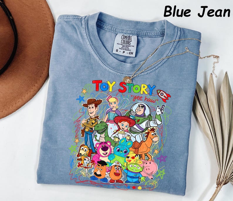 May include: Blue jean colored t-shirt featuring a colorful graphic of Toy Story characters. The text "Toy Story" and "yee haw" are displayed on the front. The shirt has a crew neck and short sleeves. A gold necklace is draped across the shirt.
