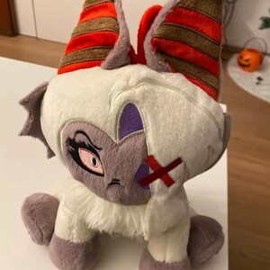 May include: A plush toy featuring a white, furry creature with large, pointed ears striped in brown and orange. The face has a purple patch over one eye, a red embroidered cross over the other, and a single visible eye with a pink iris.