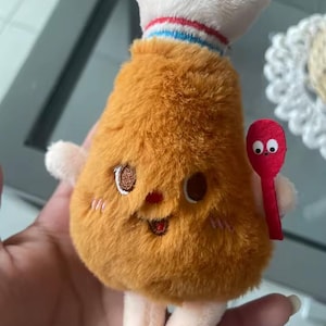 May include: A plush toy shaped like a fried chicken drumstick with golden-brown fur, a white bone top with red and blue stripes, embroidered facial features, and a small red spoon accessory held on the side with two googly eyes on the handle.