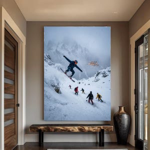 Könnte beinhalten: Ein großer gerahmter Druck einer verschneiten Berglandschaft mit Snowboardern und Skifahrern. Ein Snowboarder befindet sich in der Luft, während andere die Piste hinunterfahren. Der Hintergrund zeigt eine Bergkette und ein Gebäude. Der Druck wird über einer Holzbank angezeigt.