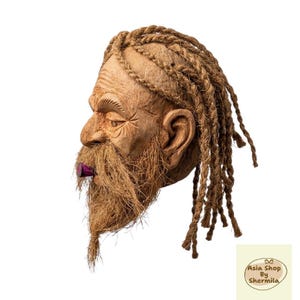 Puede incluir: Una escultura de cáscara de coco con forma de cabeza humana con una barba fibrosa marrón texturizada y cabello peinado en largas trenzas. El rostro presenta detalles tallados alrededor de los ojos y un pequeño objeto cilíndrico de color morado.