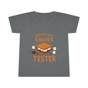 May include: A grey toddler t-shirt with the words "OFFICIAL S'MORE TESTER" in orange. The graphic features a s'more with marshmallows and chocolate.