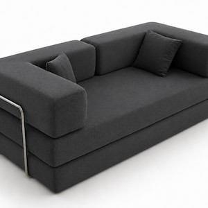 May include: A dark grey upholstered sofa featuring a low-profile rectangular design with thick, blocky armrests and backrests. The piece includes two matching square throw pillows and is supported by a visible, thin, silver-toned metal frame.