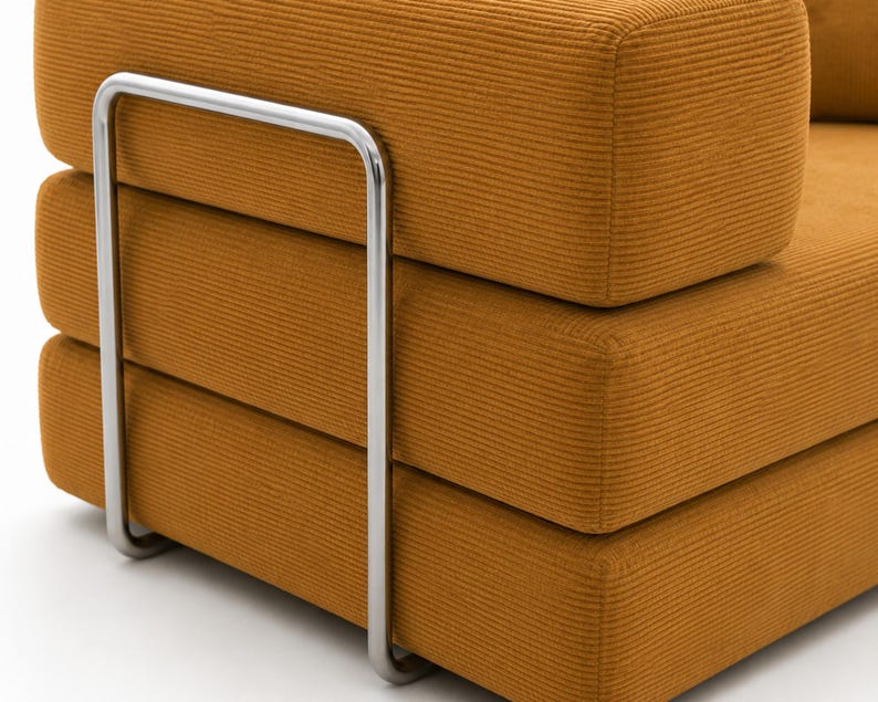 May include: A mustard yellow sofa section featuring a textured corduroy fabric upholstery. The piece is supported by a prominent, U-shaped polished chrome metal frame that wraps around the side of the thick, stacked rectangular cushions.