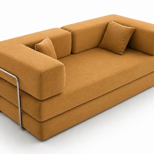 May include: A mustard yellow sofa with a textured fabric finish, featuring a low-profile design with thick, blocky armrests and backrests. The piece is supported by a visible silver-toned metal frame on the side and includes two matching square pillows.