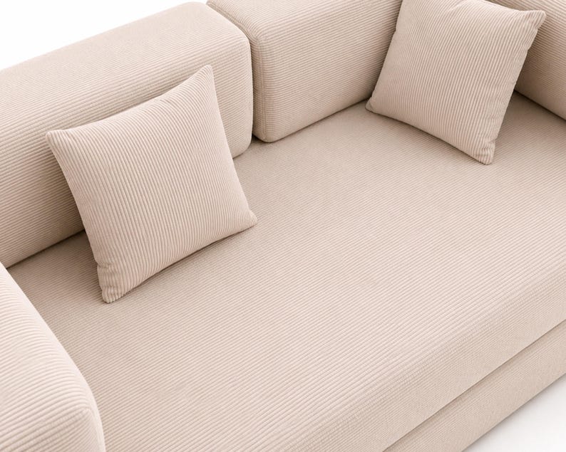 May include: A beige corduroy sofa featuring a textured, ribbed fabric surface. The furniture includes two matching square throw pillows placed on the seat, with thick, padded armrests and a continuous, uniform fabric finish across all visible parts.