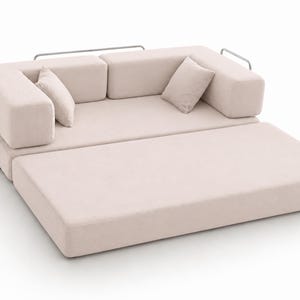 May include: A light beige modular sofa bed featuring a low-profile design with a metal frame, two square back cushions, and two smaller throw pillows. The base extends outward to create a flat sleeping surface, upholstered in a solid light fabric.