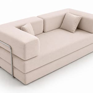 May include: A beige sofa with a textured fabric finish, featuring a low-profile design, thick rectangular armrests, and two matching square throw pillows. The structure is supported by a visible, thin, silver-toned metal frame on the side.