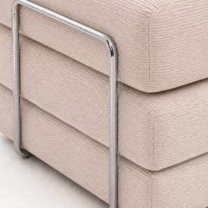 May include: A beige ottoman featuring a textured corduroy fabric upholstery and a polished chrome metal frame that wraps around the sides. The piece consists of three stacked, rounded rectangular cushions with visible horizontal seams.