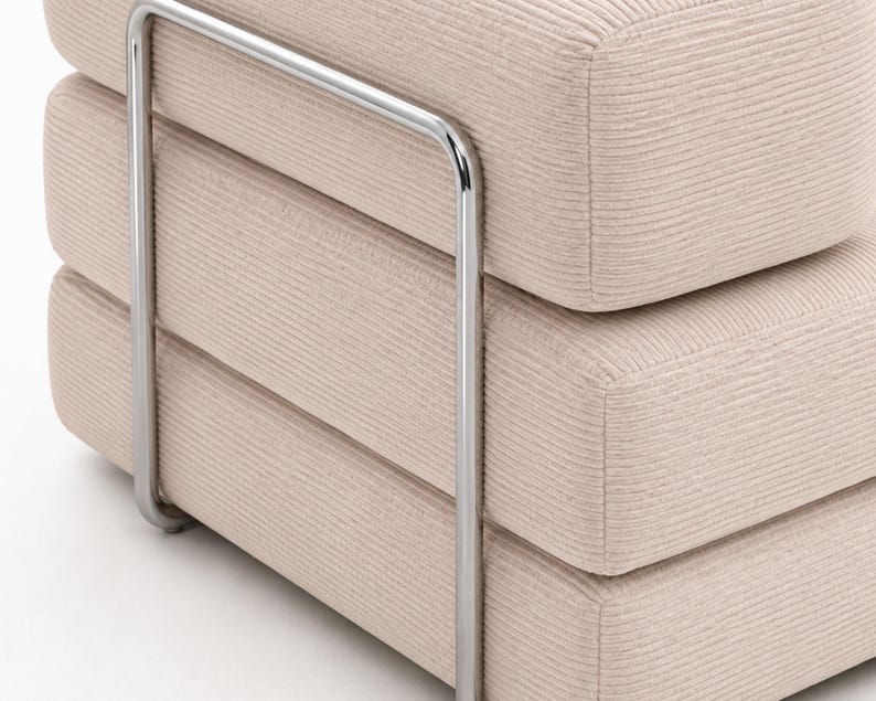 May include: A beige ottoman featuring a textured corduroy fabric upholstery and a polished chrome metal frame that wraps around the sides. The piece consists of three stacked, rounded rectangular cushions with visible horizontal seams.