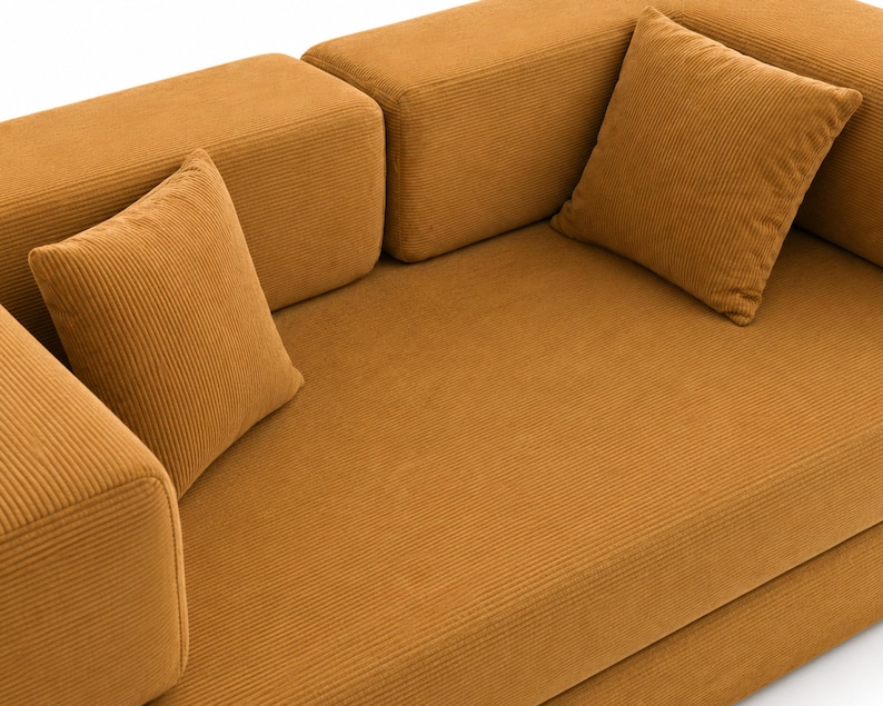 May include: A mustard yellow corduroy sofa featuring a textured ribbed fabric finish, thick rectangular armrests, and two matching square throw pillows placed on the seat. The furniture has a clean, blocky design with a uniform golden-brown color.
