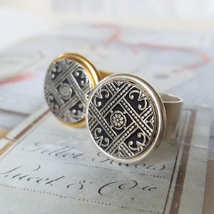 May include: Two silver rings with black and silver patterned designs. The rings are on a white surface with black writing.