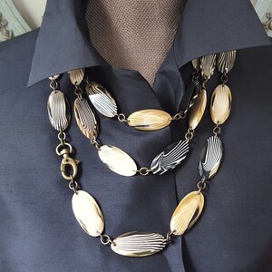 May include: A three-strand necklace with oval-shaped beads in shades of cream, black, and tan. The beads have a striped pattern and are connected by gold-colored chain links. The necklace is displayed on a dark blue collared shirt.