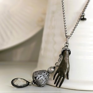 May include: A silver-toned necklace featuring a pendant shaped like a hand. The hand pendant is detailed with a textured finish and is connected to a chain. Additional charms include a faceted teardrop, a filigree heart, and a small bead.