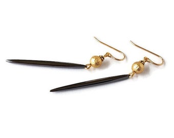 May include: A pair of drop earrings featuring long, slender, dark gray spikes suspended from gold-toned ear wires. Each earring includes a small, round, gold-colored bead with a textured, hammered finish positioned above the spike.