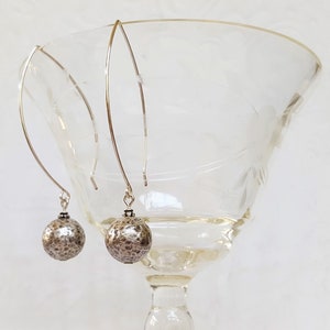May include: A pair of silver dangle earrings with textured silver spheres. The earrings are hanging from a glass goblet.
