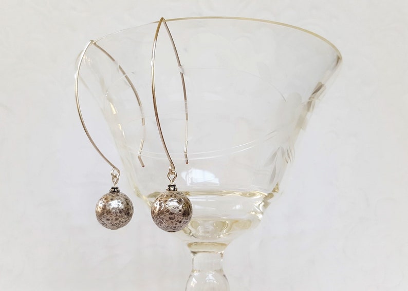 May include: A pair of silver dangle earrings with textured silver spheres. The earrings are hanging from a glass goblet.