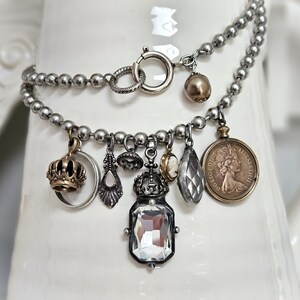 May include: A silver charm bracelet featuring various pendants. Charms include a gold crown, a clear crystal drop, a cameo, and a coin. The bracelet is shown against a white backdrop.