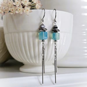 May include: A pair of dangle earrings with a light blue, cube-shaped bead, silver-colored chain, and a silver-colored bar. The earrings have a silver-colored hook and are displayed in front of a white bowl with flowers.
