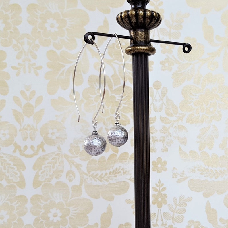 May include: A pair of silver dangle earrings with hammered round silver beads. The earrings are hanging from a silver stand.
