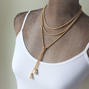May include: A gold chain necklace with a long, dangling chain that has two pearl accents at the end. The necklace is layered with two shorter chains.