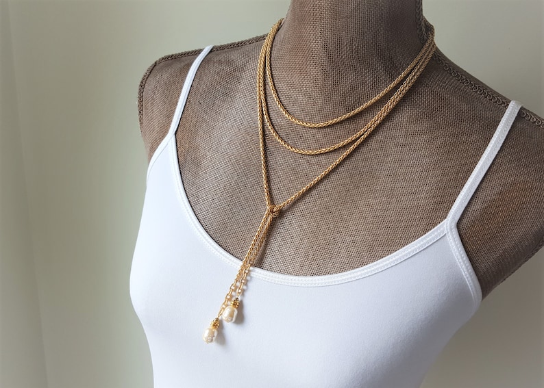 May include: A gold chain necklace with a long, dangling chain that has two pearl accents at the end. The necklace is layered with two shorter chains.