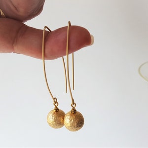 May include: A pair of gold dangle earrings with hammered ball charms. The earrings are made of brass and have a simple, elegant design.