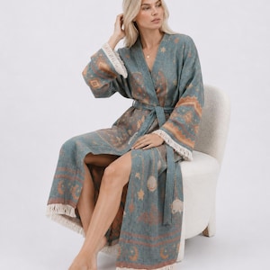 May include: A long-sleeved teal robe featuring a woven pattern of celestial moons, stars, and geometric shapes in muted orange and cream. The garment includes a matching tie belt at the waist and cream-colored fringe trim along the sleeve cuffs and hem.