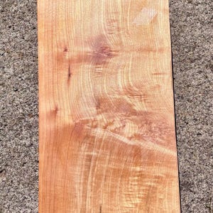 May include: A rectangular wooden board with a light brown, natural finish. The surface displays a prominent, swirling grain pattern with visible knots and organic wood textures throughout the piece, showcasing a smooth, flat, and polished appearance.