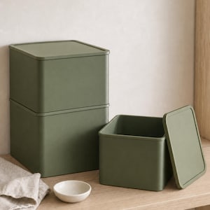 Può includere: Un set di tre contenitori quadrati color verde oliva opaco con coperchi. Due contenitori sono impilati verticalmente, mentre il terzo si trova sulla destra con il coperchio rimosso accanto. Una piccola ciotola bianca è in primo piano.